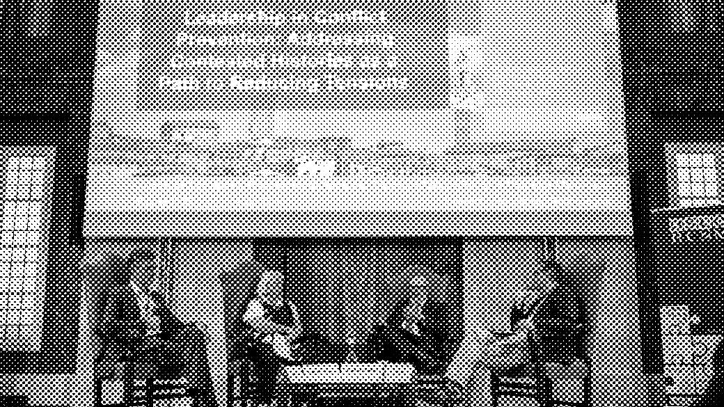 Panel discussion with four people seated, large screen and banners in the background. Casual setting, formal attire.