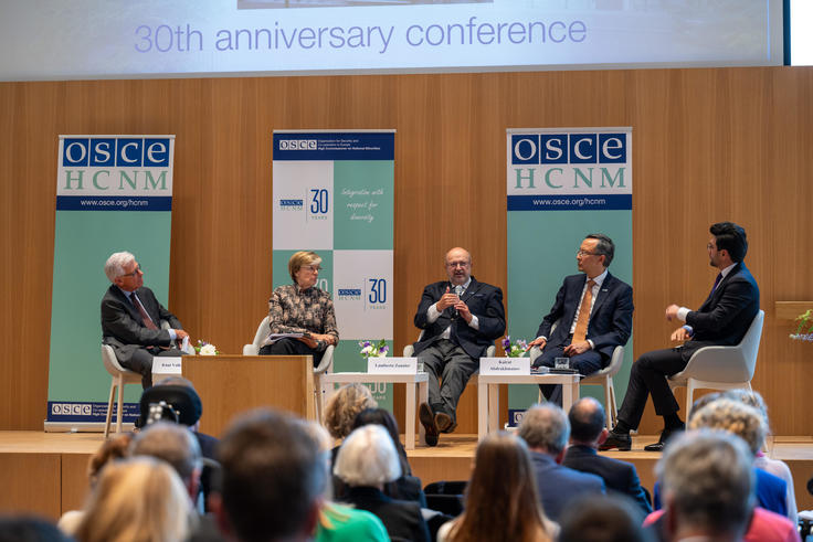 Panel of five people discussing on stage at a conference, seated with microphones, facing an audience.