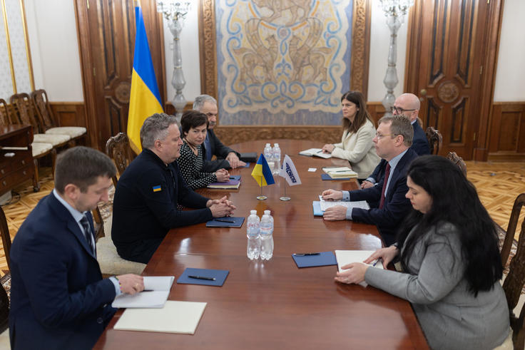 Meeting in a formal room, two groups of people sit across a table with Ukrainian and EU flags in the background.