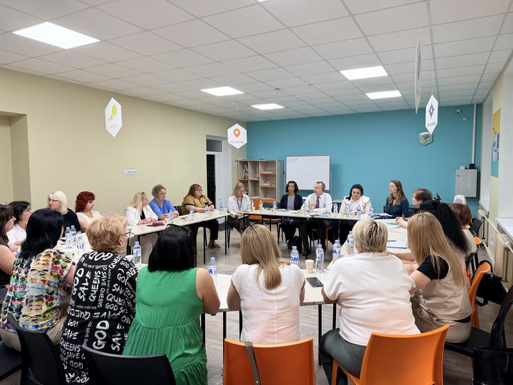 A group of people sit around a long table in a brightly lit meeting room, engaged in discussion.
