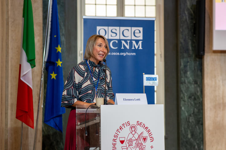 Woman speaking at a podium with flags in the background, including EU and Italian flags.