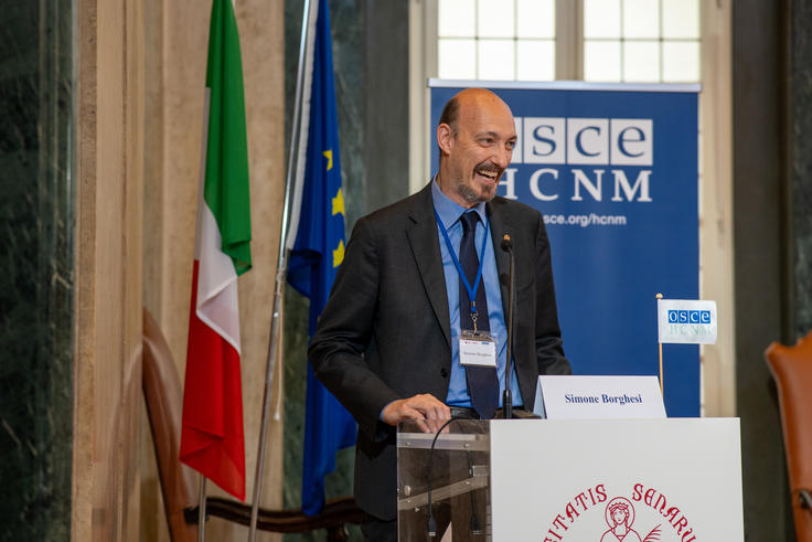 Man speaking at a podium with flags and a blue conference banner in the background.