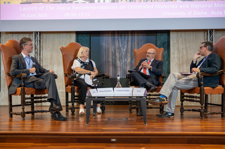 Four people seated on a stage in a panel discussion, wearing formal attire, with a table and chairs on a wooden floor.