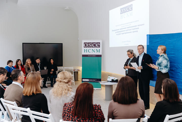 Presentation in a modern conference room with three speakers addressing an audience, seated in white chairs.
