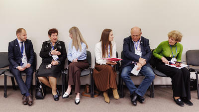 Seven people sitting in a row, engaged in conversation, wearing business attire in a meeting room.
