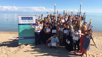 Group of people celebrating on a sunny beach, holding certificates with a scenic lake and mountains in the background.