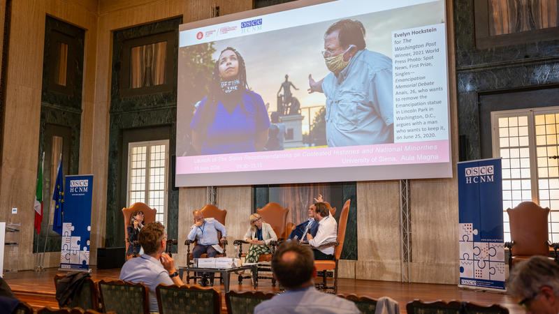 Panel discussion in a large hall with people seated, a screen displaying two individuals, and posters on the walls.