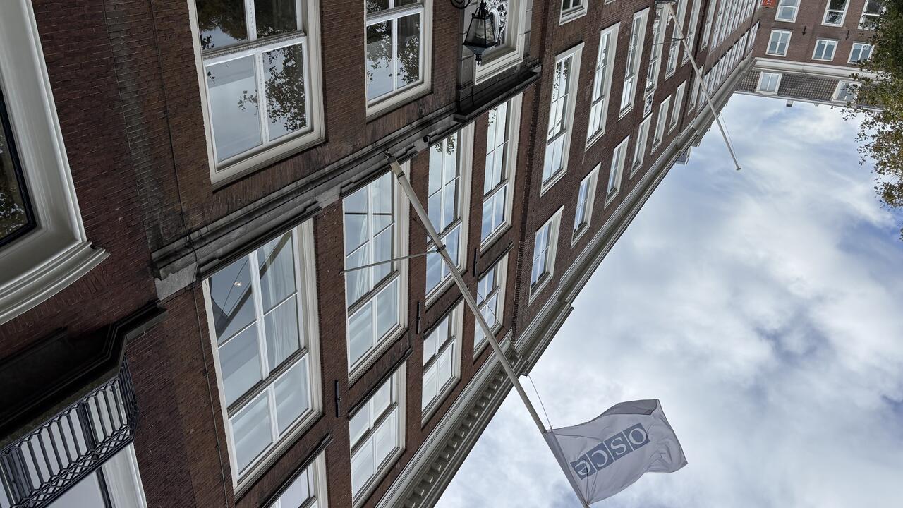 Red brick building with many windows, a white flag with text, and a cloudy blue sky above.