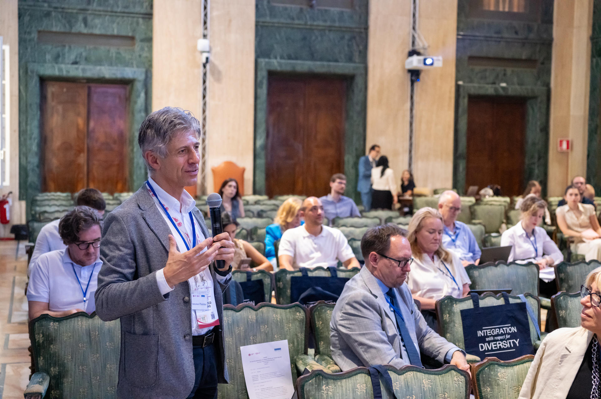 Man speaking with a microphone in a conference room, audience listening attentively.