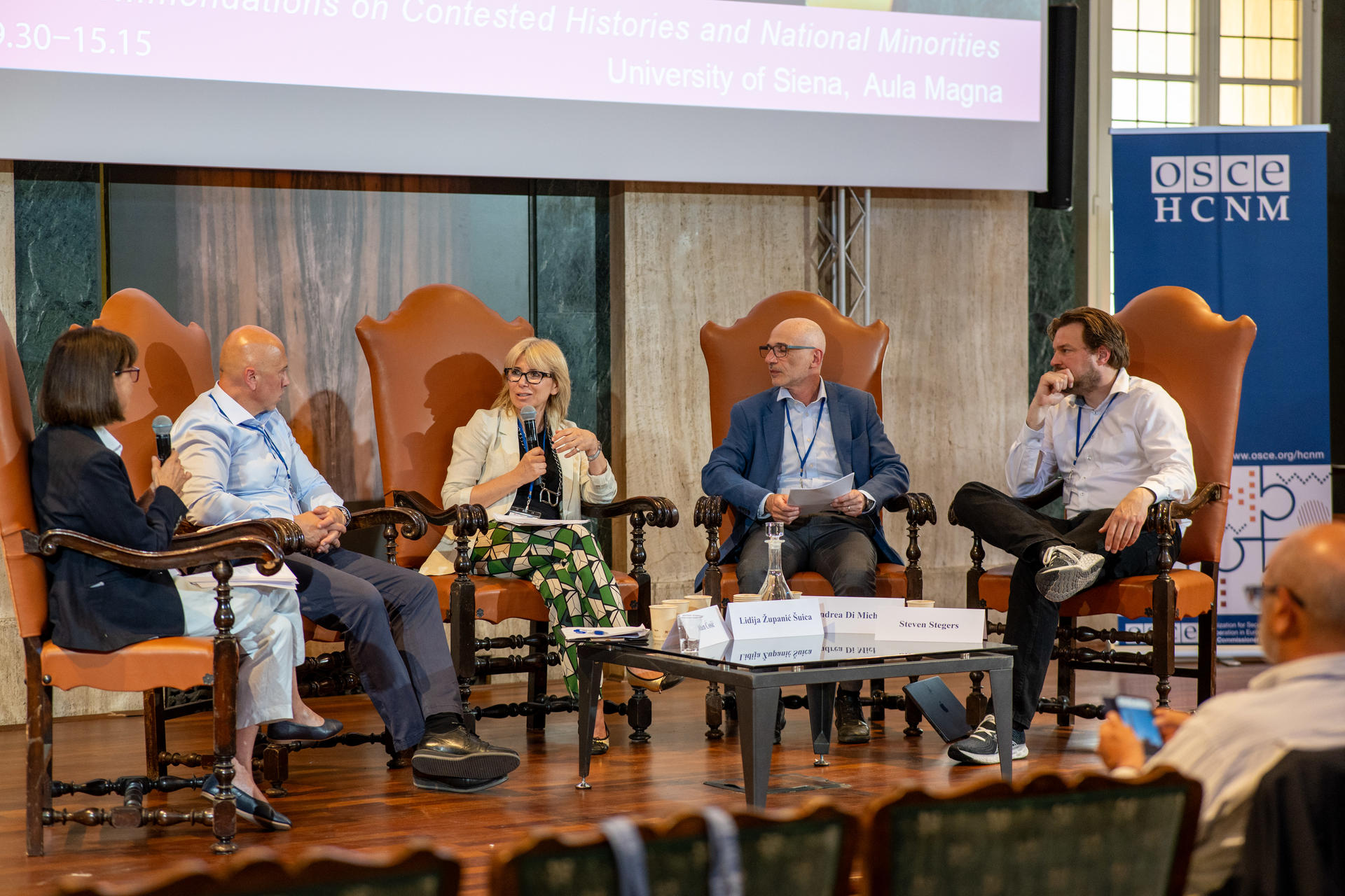 Panel discussion with five people seated in large chairs, speaking and listening intently in a conference setting.