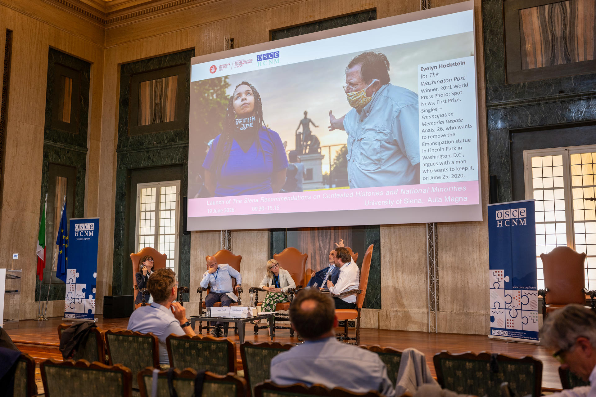 Panel discussion in a large hall with people seated, a screen displaying two individuals, and posters on the walls.